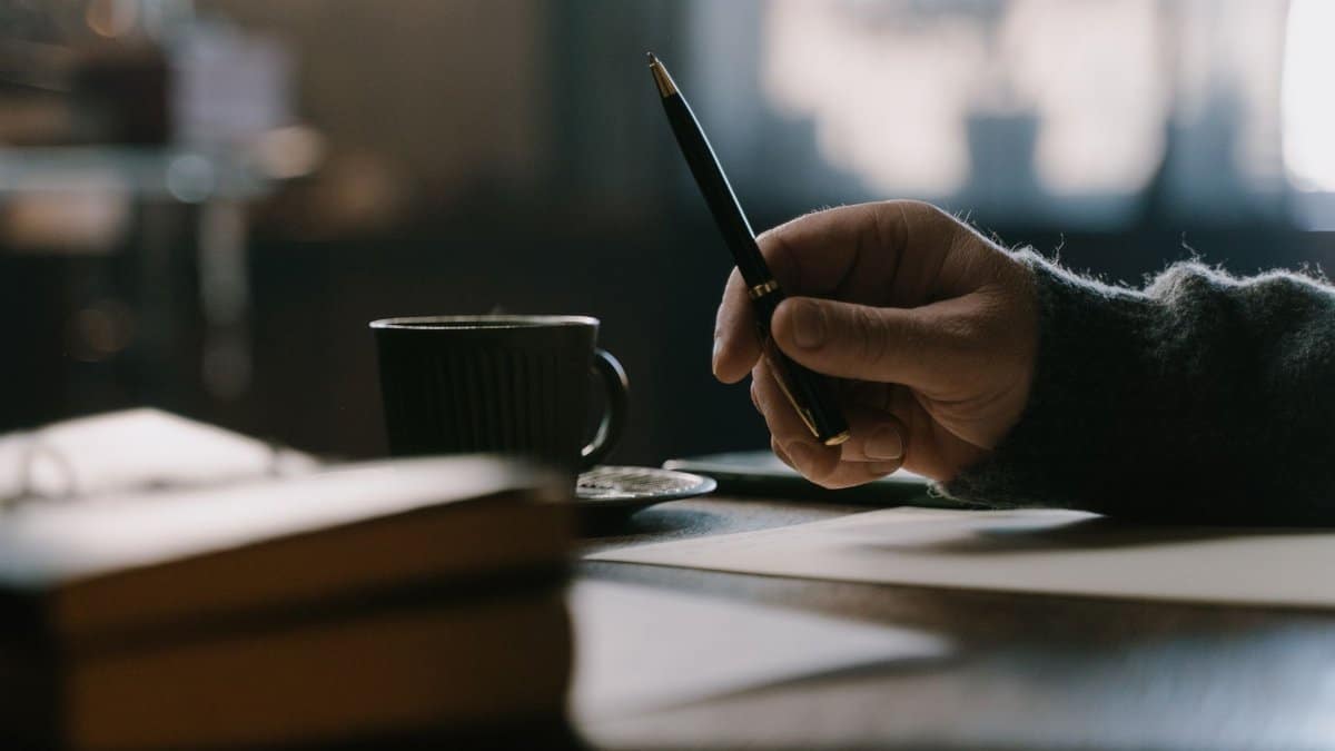Hand holding pen at a table with a cup and books, evoking a cozy ambiance.