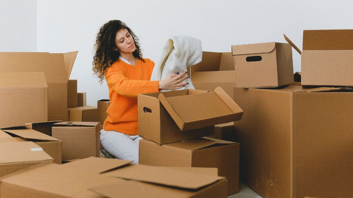 Calm young relocating woman with curly dark hair in stylish sweater packing belongings into pile of cardboard boxes sitting on floor before moving into new apartment