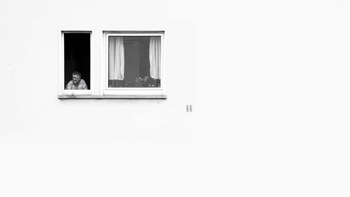 A man looks pensively out of an open apartment window, captured in monochrome.