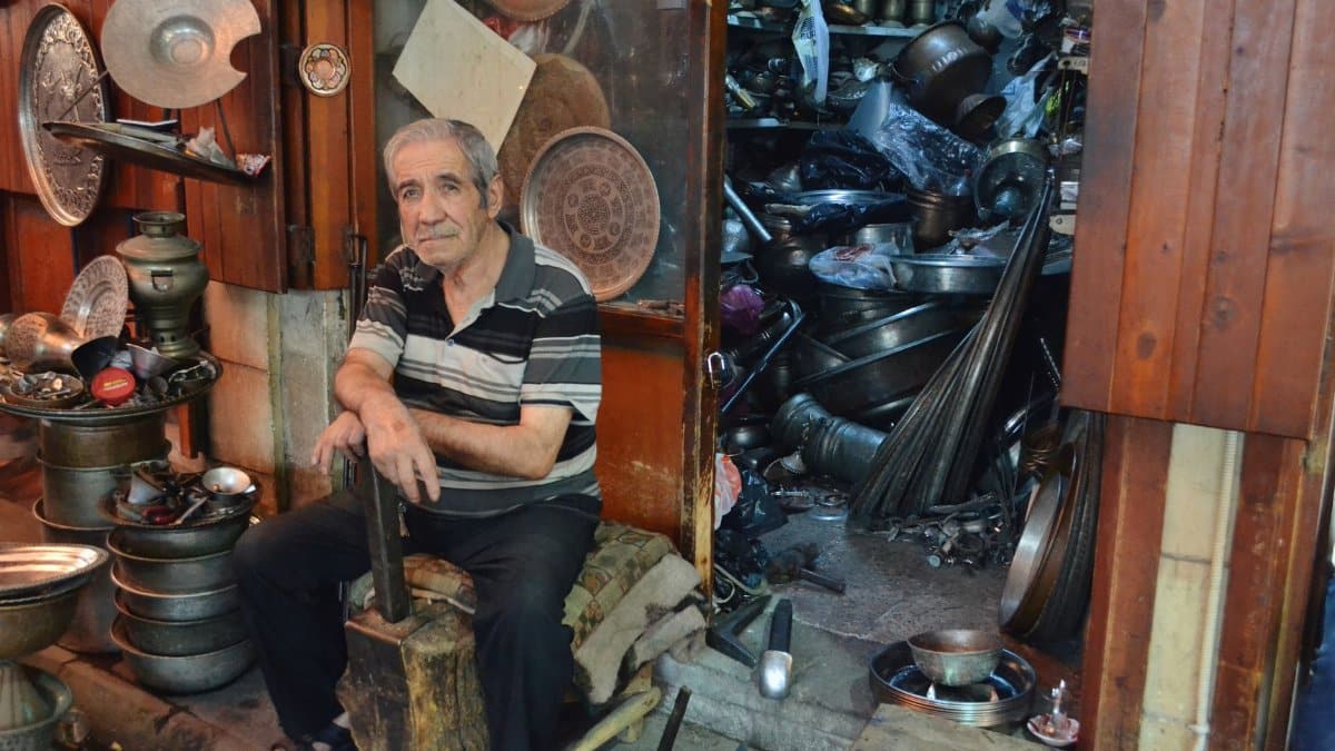 Elderly man sitting in a cluttered metal workshop surrounded by metal objects and tools.