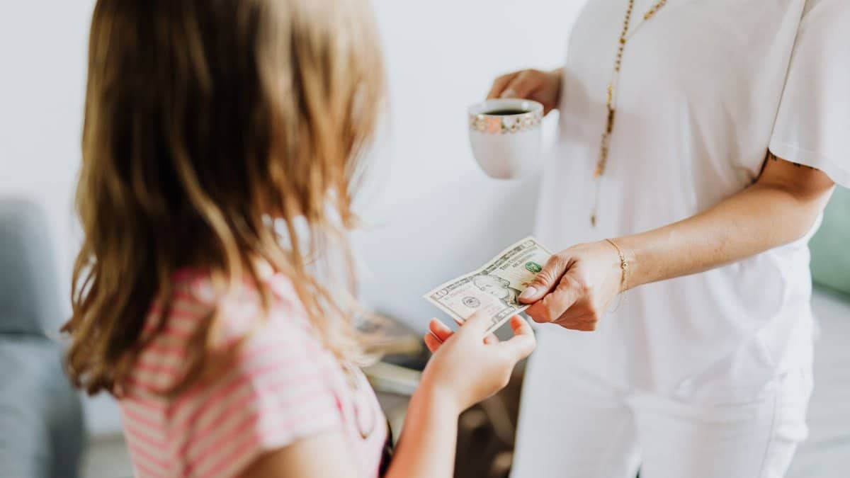 A woman hands a dollar bill to her daughter while holding a coffee cup, indoors.
