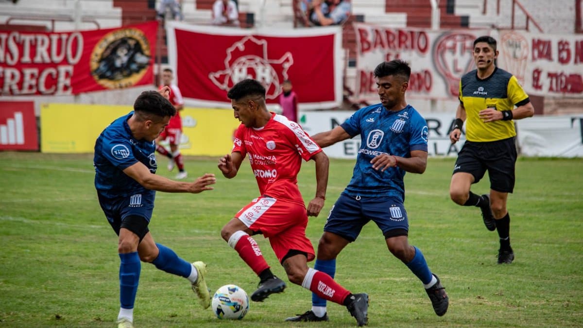 Soccer players in action during a competitive match on a grass field with spectators in the stands.