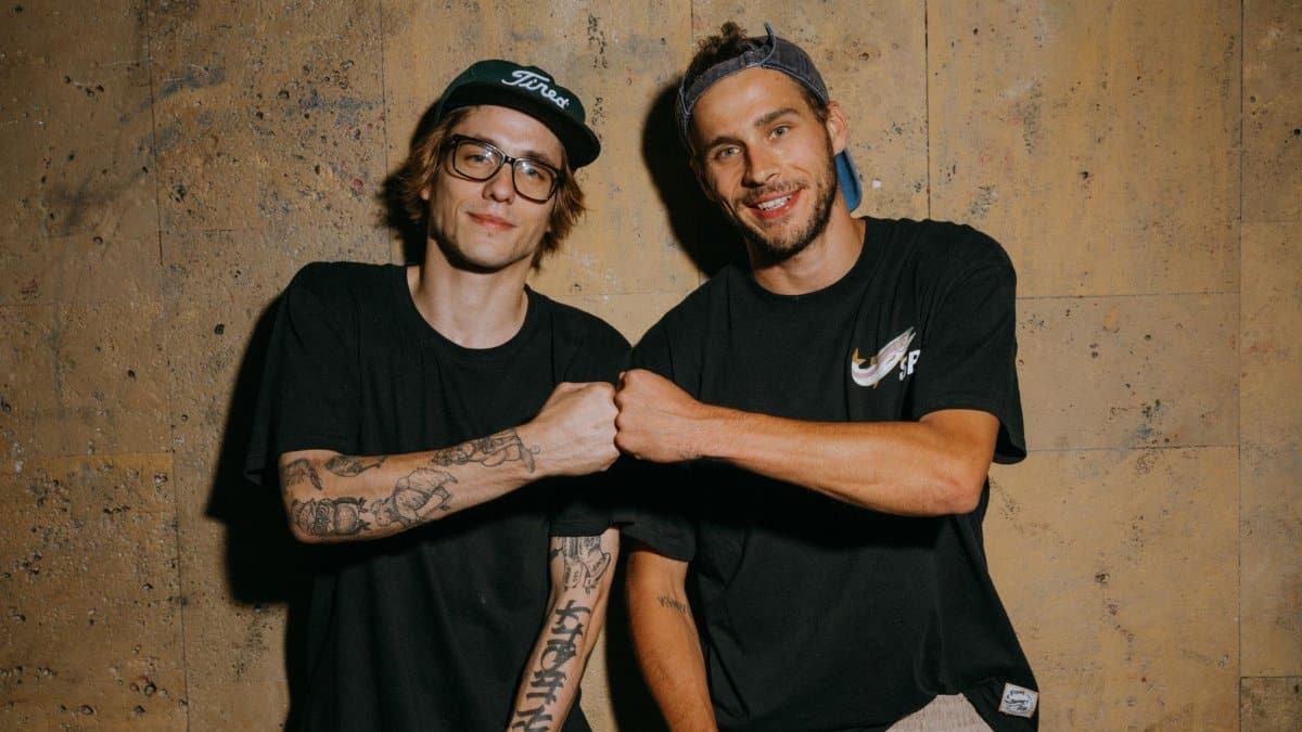 Two young men in casual clothing smiling and fist bumping against a textured wall indoors.