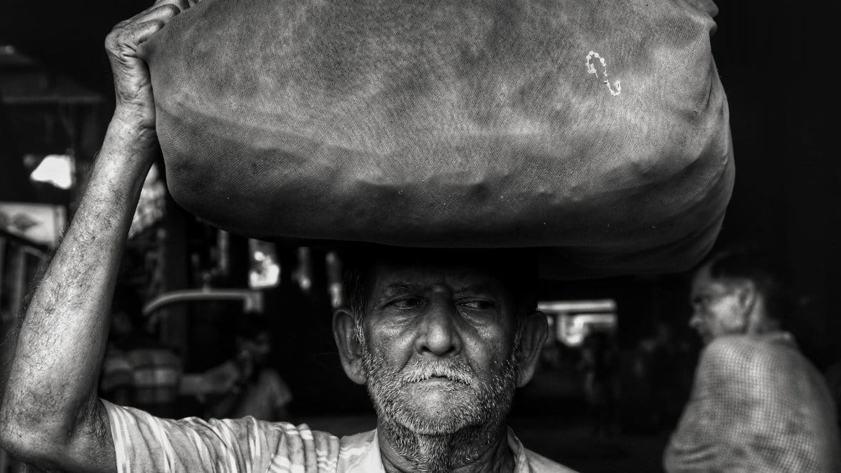 A hard-working elderly man carries a large sack on his head in grayscale.