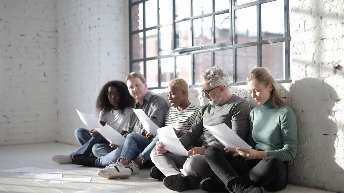 Group of contemporary diverse coworkers in casual wear with papers sitting with crossed legs on ceramic floor near fenced window in afternoon