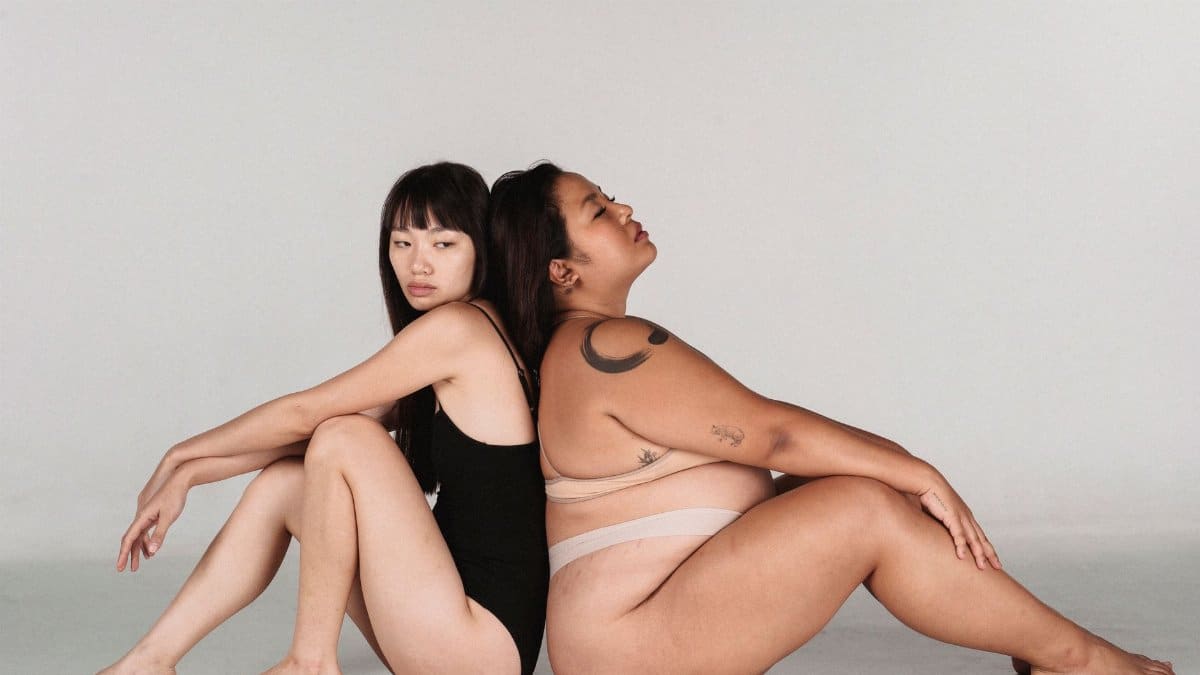 Side view of Asian women in underwear sitting back to back in studio against grey background