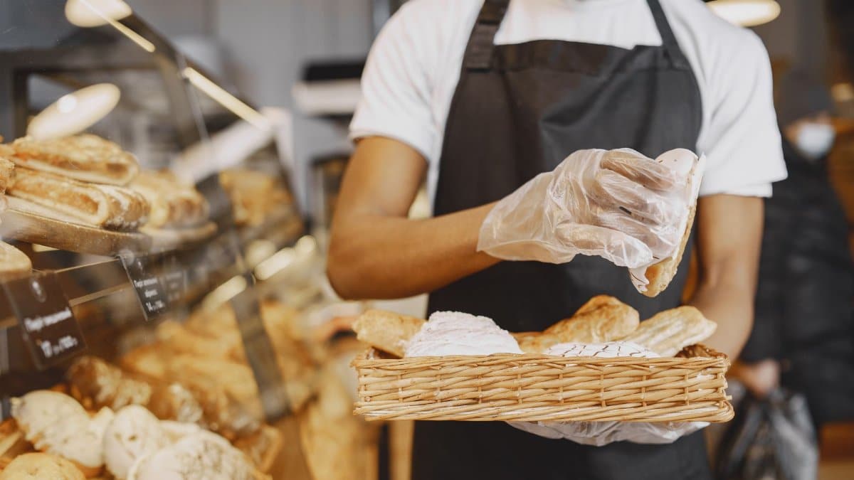 A baker wearing a mask and gloves prepares fresh pastries in a bakery environment.