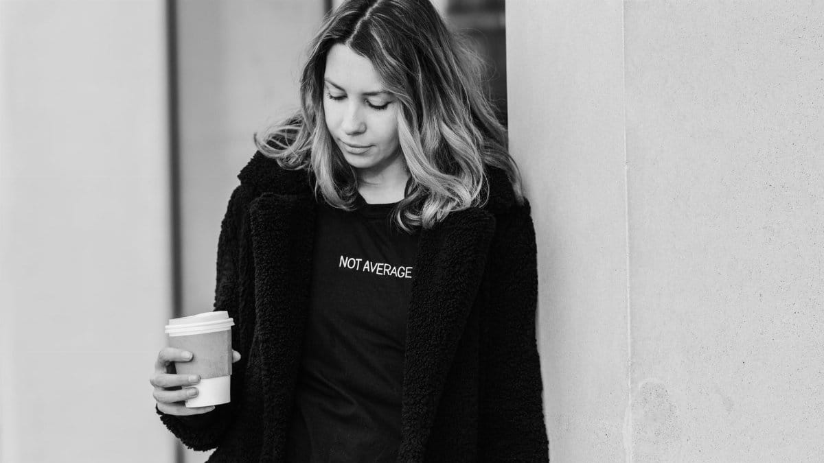 Black and white of adult tender female in outerwear with disposable glass of hot drink looking down in city