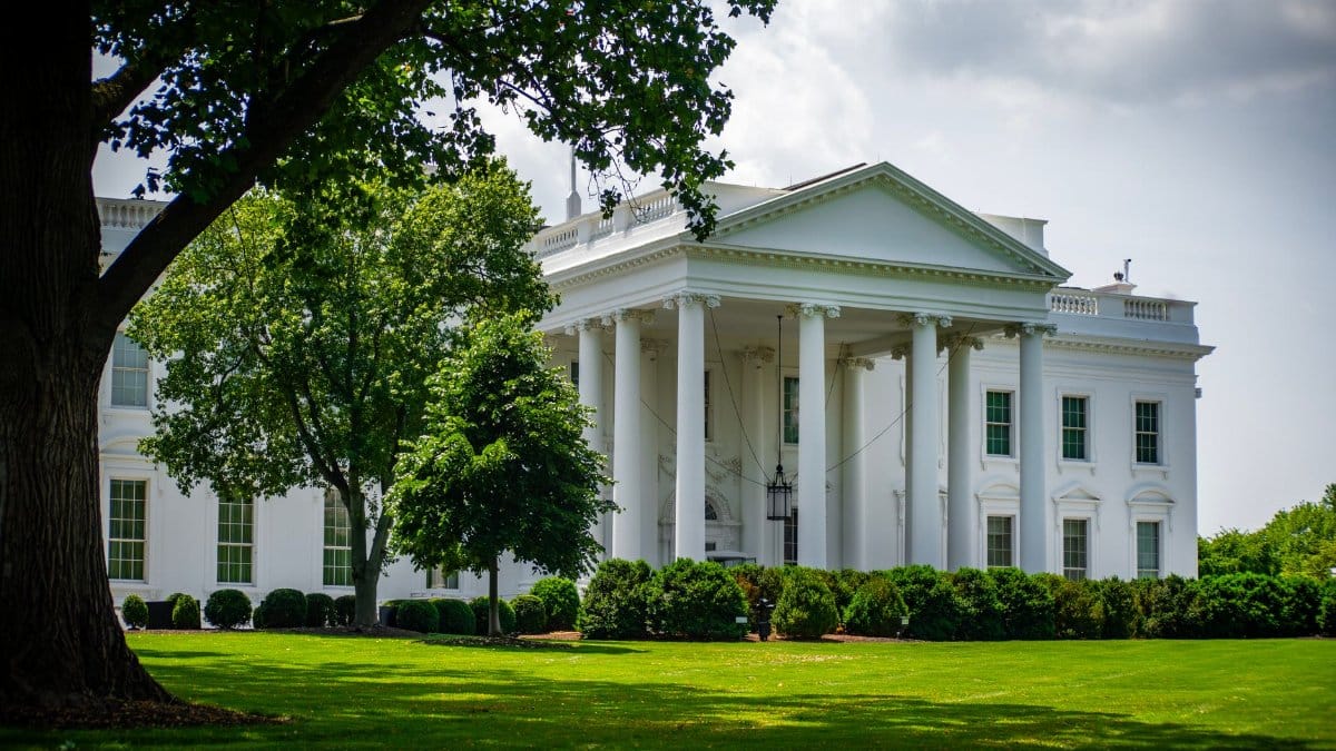 The White House framed by trees and greenery, in Washington, D.C., under a bright sky.
