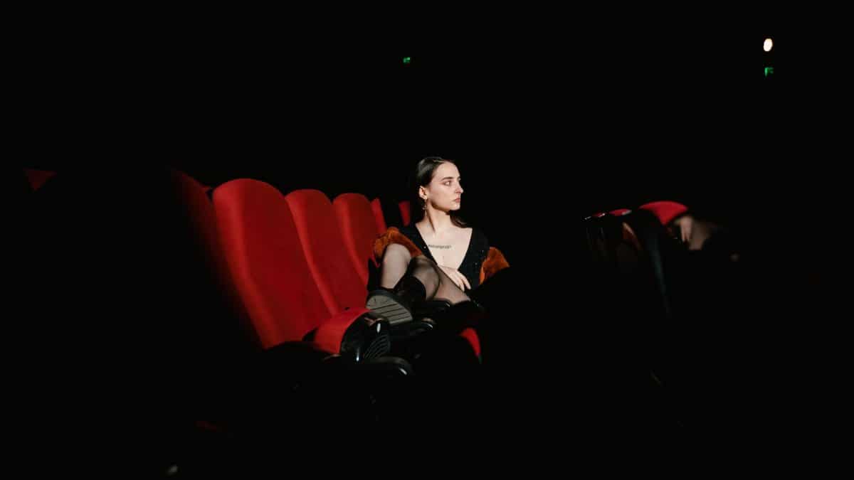 A lone woman sits in an empty theater, illuminated by soft light on red seats, creating a dramatic feel.