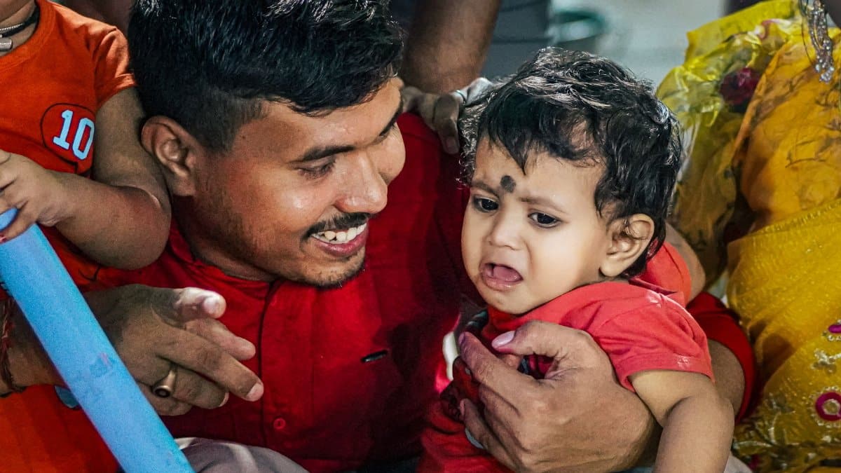 A father lovingly comforts a crying baby at a lively family gathering indoors.