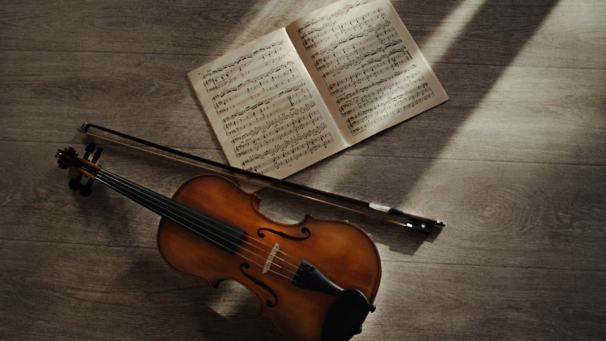 Artistic composition of a violin, sheet music, and bow in warm sunlight on a wooden floor.