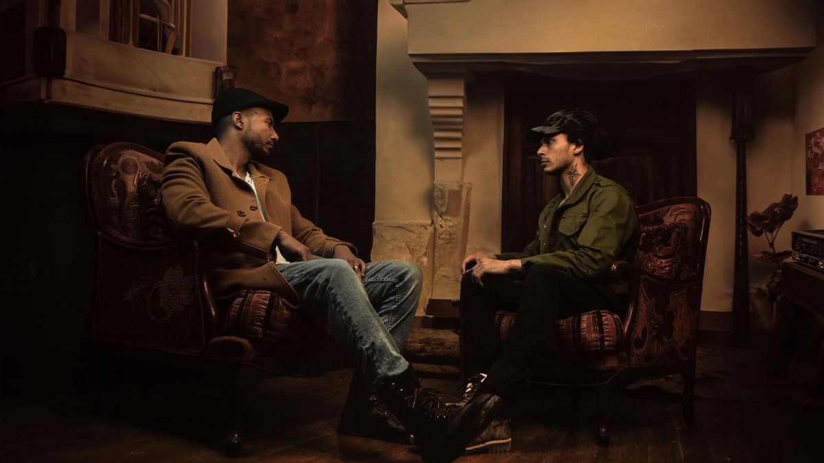 Two men in fashionable attire sit and converse in a dimly lit room in Los Angeles.