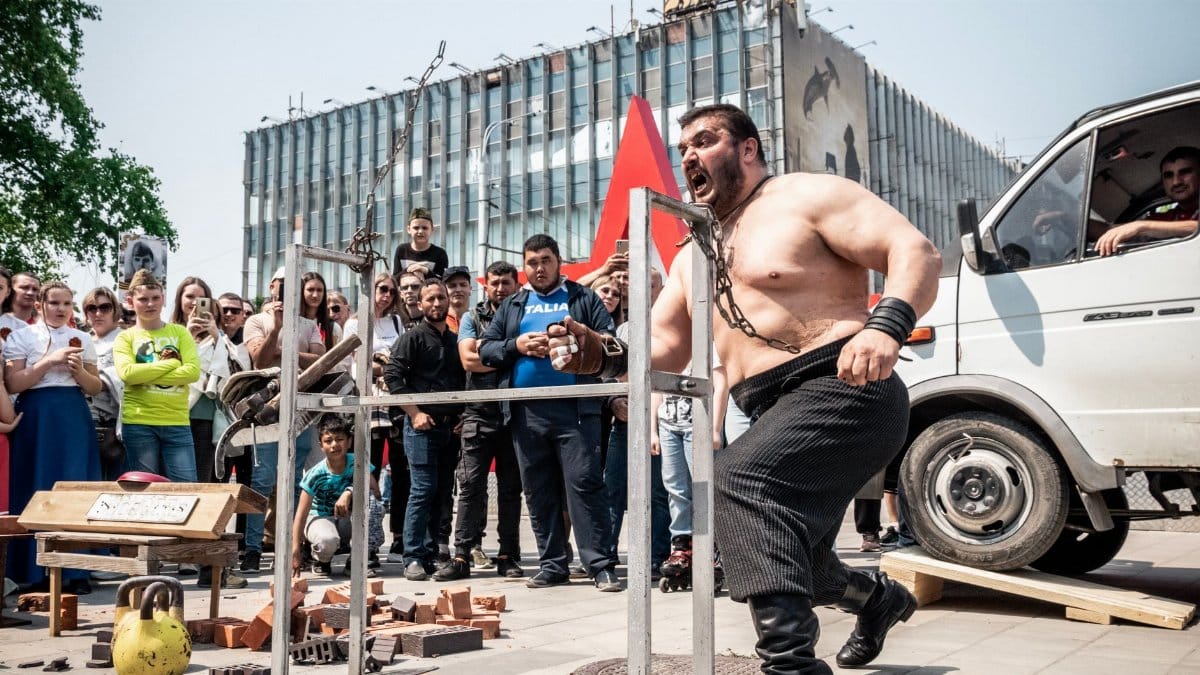 A strongman demonstrates his strength in an outdoor performance, captivating a crowd.