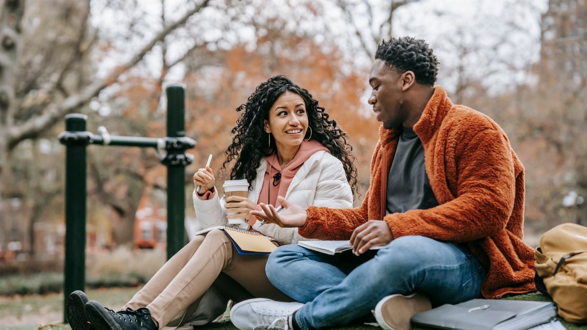 Full body of cheerful ethnic woman with takeaway coffee and black man with copybooks having conversation while preparing homework together on street