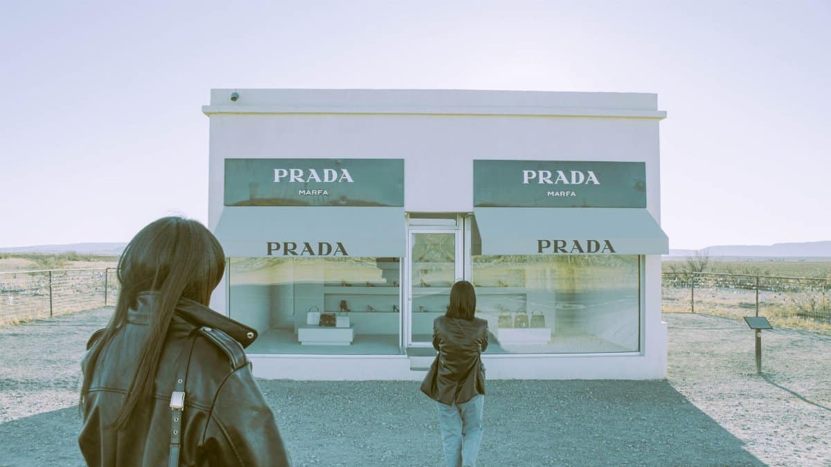 A woman stands before the famous Prada Marfa installation in Valentine, Texas under a bright blue sky.