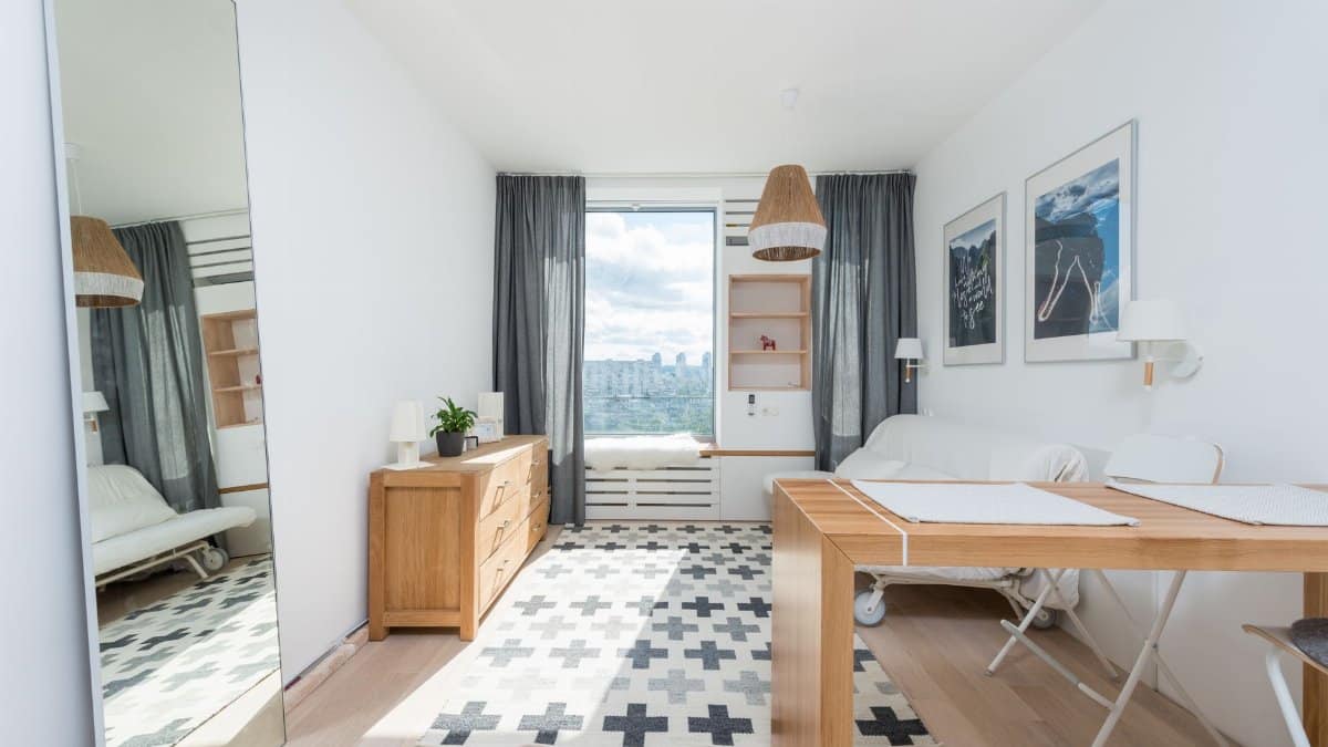 Wooden table and drawer placed in room with big window and wall decorated with pictures in light room with mirror