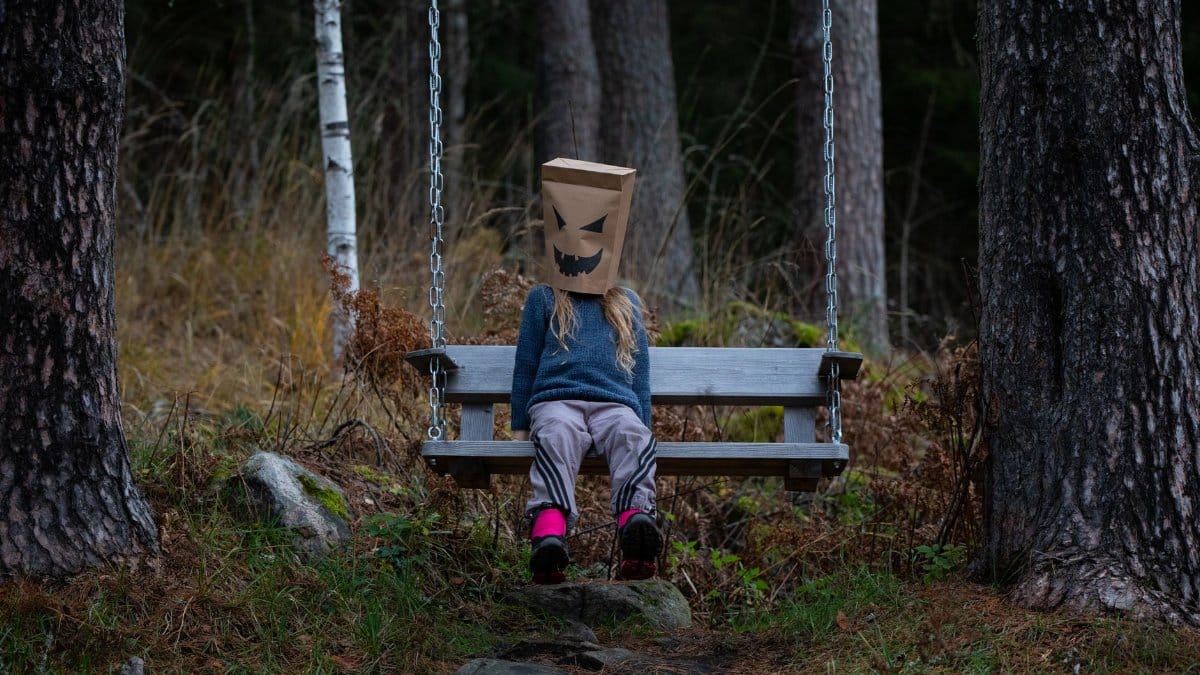 A young child wearing a playful paper bag mask swings alone in a forest setting.