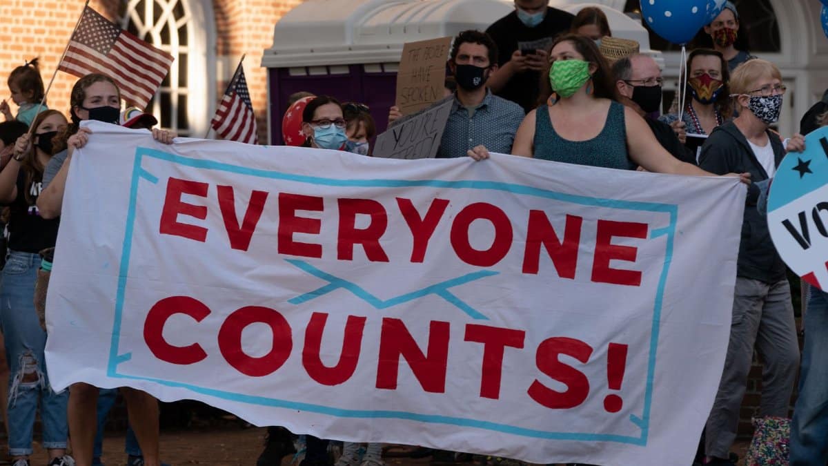 A diverse group of masked protestors holding a 'Everyone Counts' banner advocating for democratic rights.