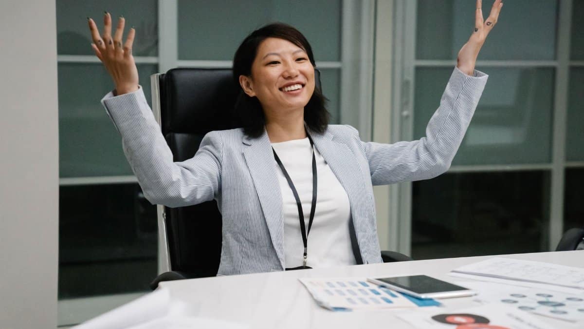 Asian businesswoman smiling and raising her hands in an office setting, celebrating success.