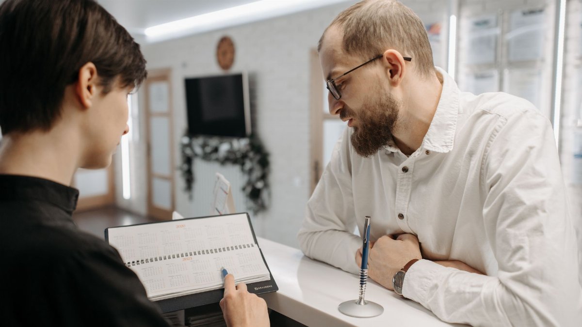 A receptionist and client converse over an appointment book at a clinic reception desk.