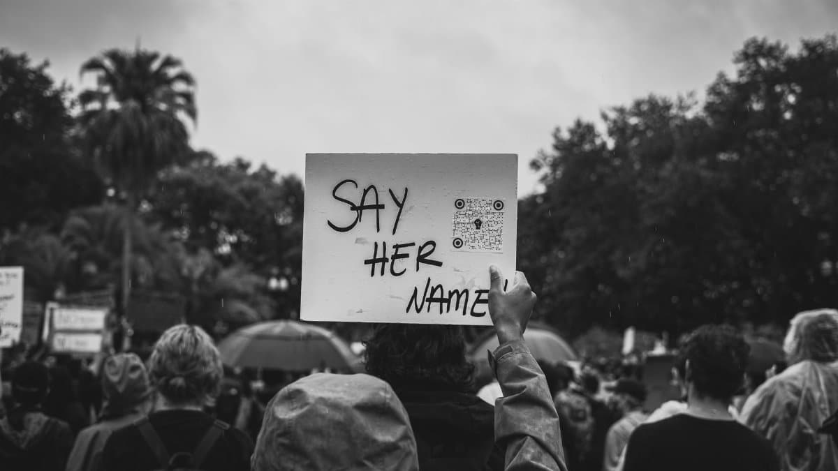 Black and white image of a protest with a 'Say Her Name' sign raised in a crowd.