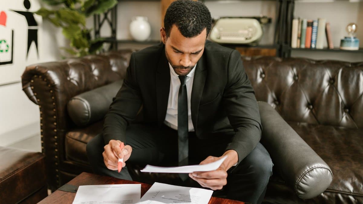 A businessman in a suit attentively reviews paperwork on a brown leather sofa.