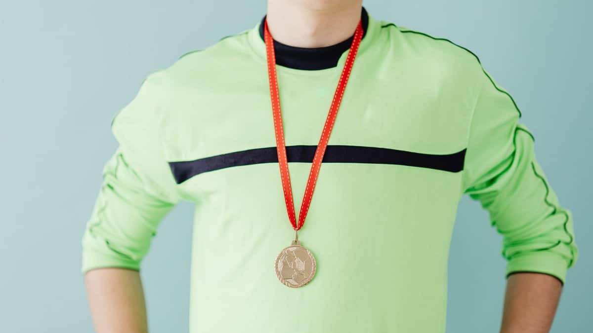 Teenager in sportswear proudly wears a gold medal with a confident pose, set against a blue background.