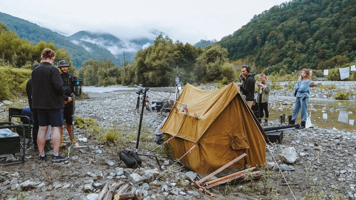 Film crew setting up camp by the river amidst forested mountains, capturing scenic landscapes.