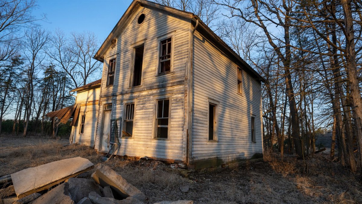An old, abandoned two-story wooden house amidst leafless trees in winter light.