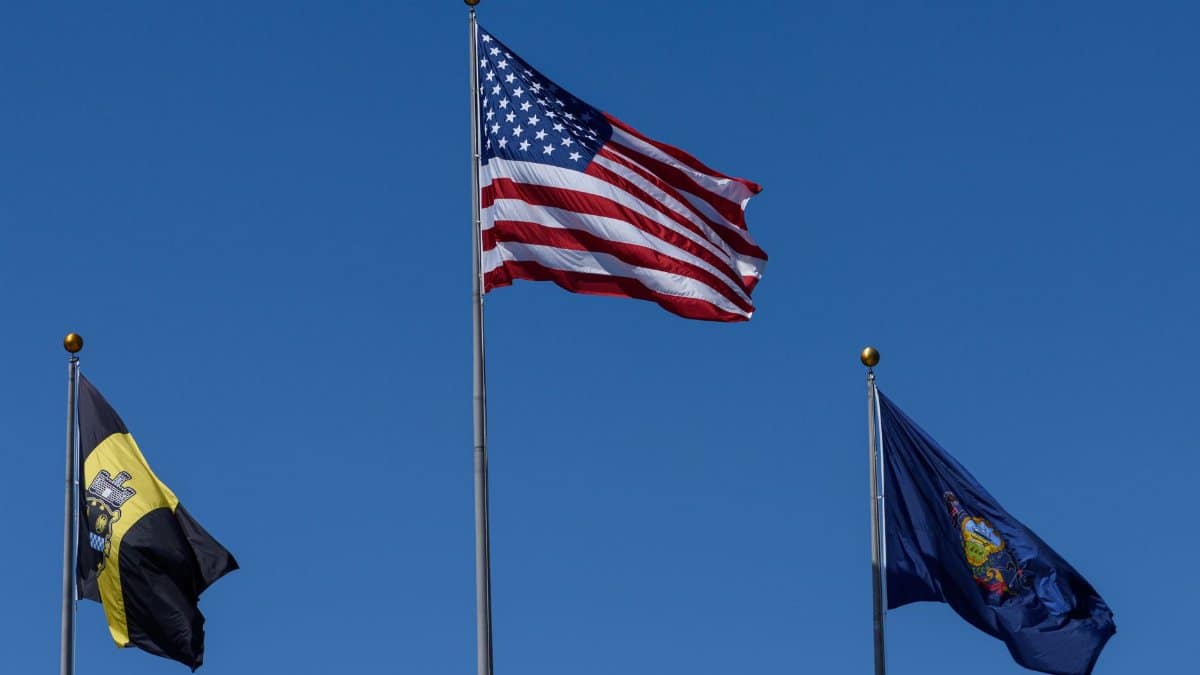 Three flags including the US flag waving against a clear blue sky.