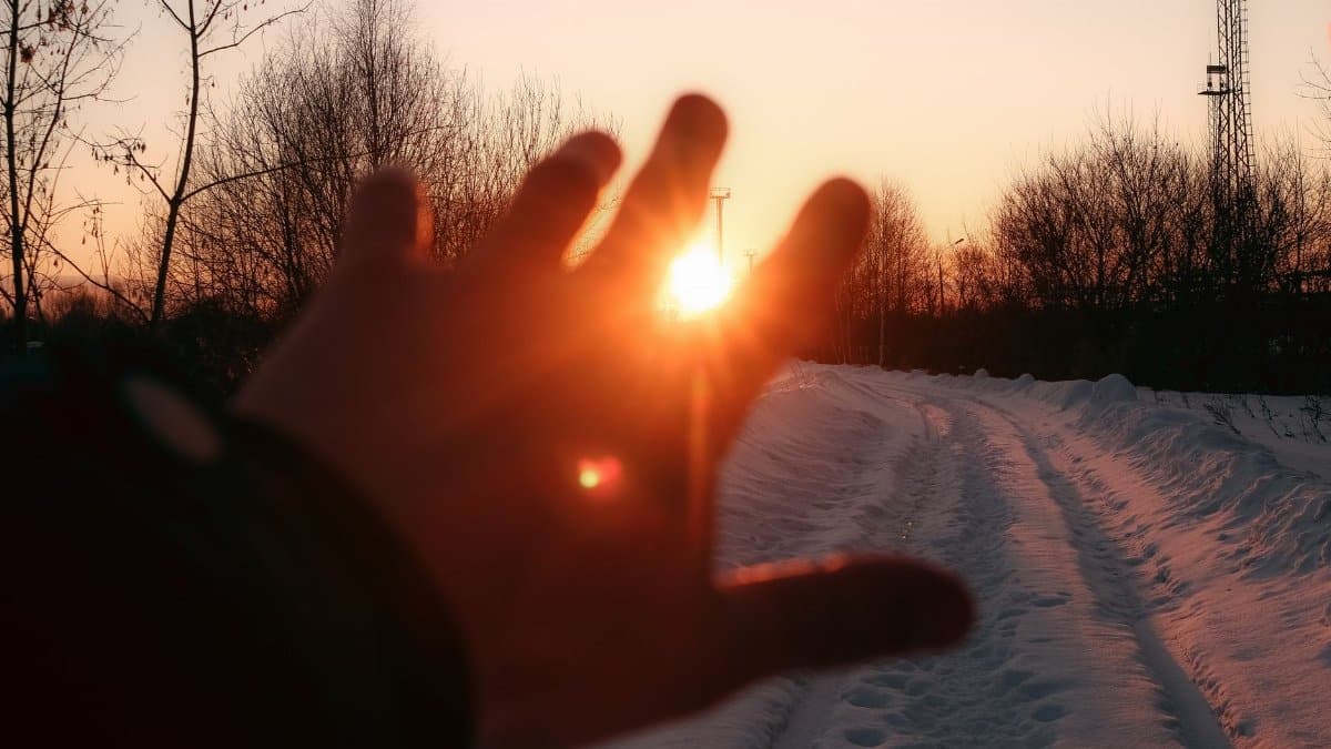 A hand reaching towards the setting sun on a snowy path, evoking warmth and tranquility.