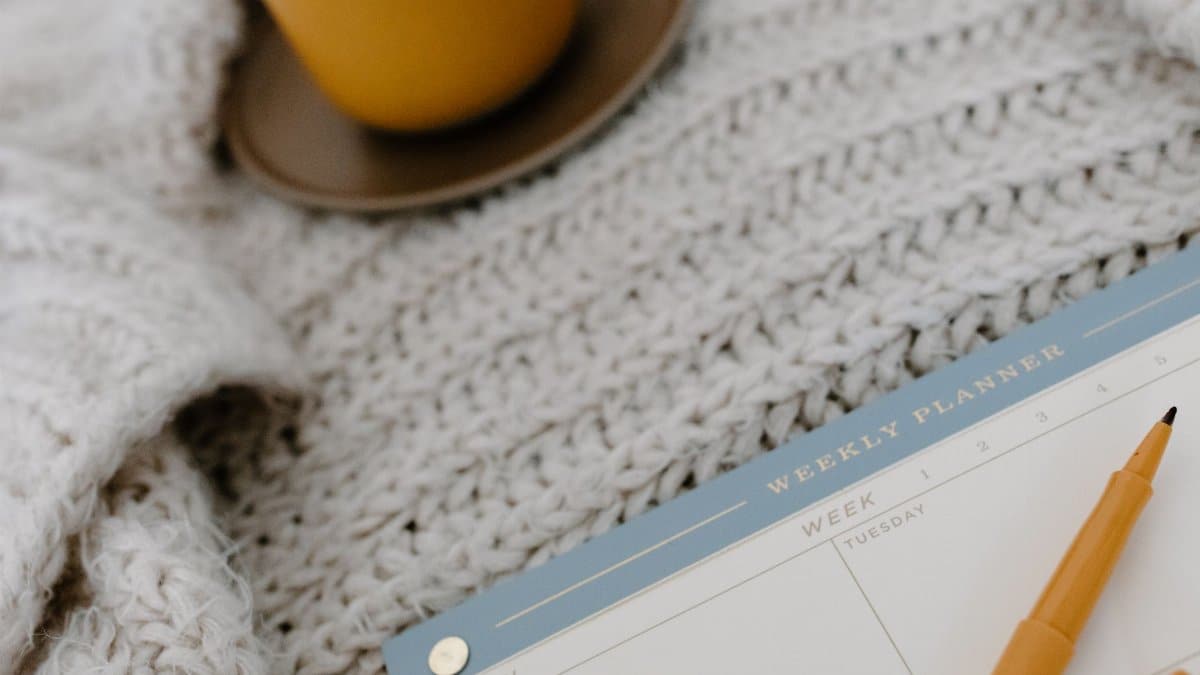 A cozy flat lay setup with a weekly planner, pen, and warm drink on a knitted blanket.