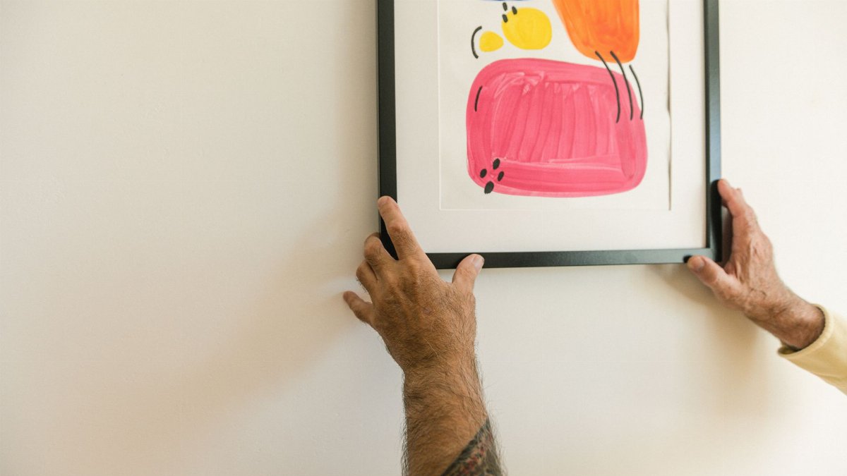 Two tattooed hands hang a colorful abstract framed artwork on a white wall indoors.