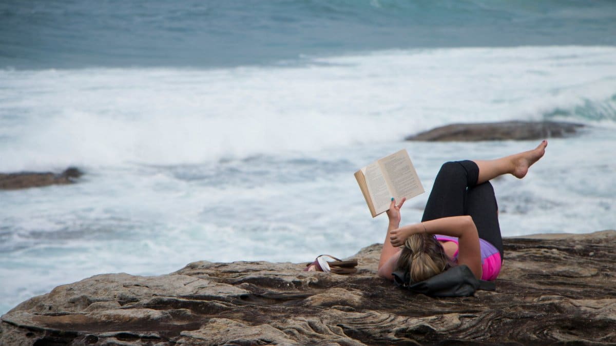 A woman enjoys reading a book while lying on rocky coast overlooking the sea.