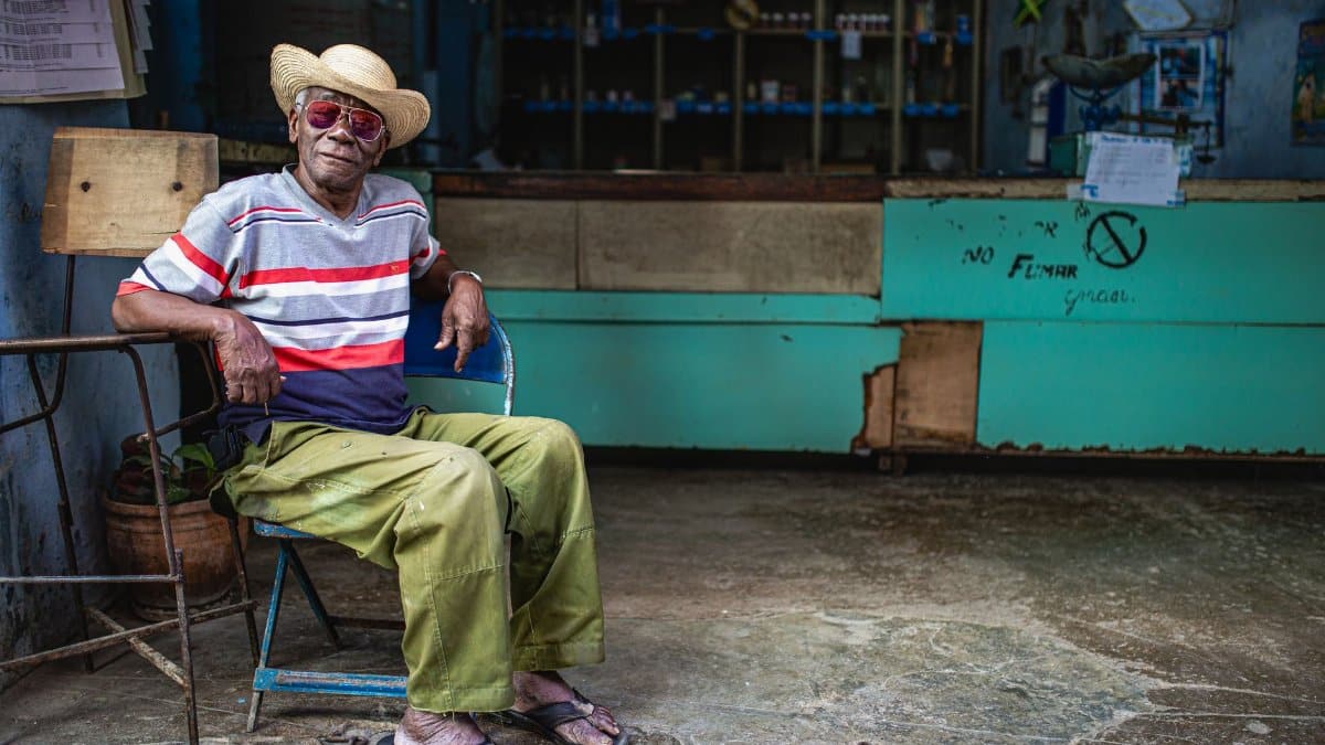 An elderly man in a hat and sunglasses sits relaxed in a rustic shop in Havana, Cuba.