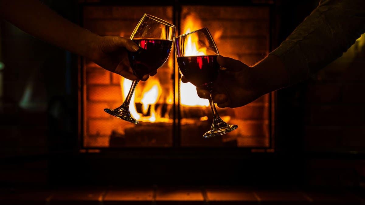 Two individuals toasting with red wine glasses beside a warm fireplace, creating a cozy ambiance.