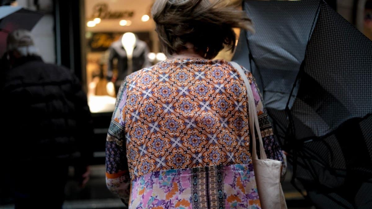 Woman with umbrella battles the wind in vibrant attire, captured candidly from behind.