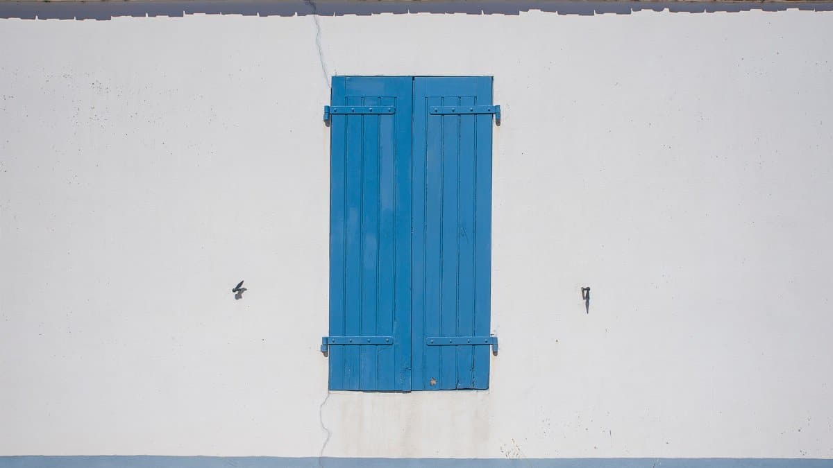 Simple minimalistic exterior with a closed blue shutter on a white wall and red roof.