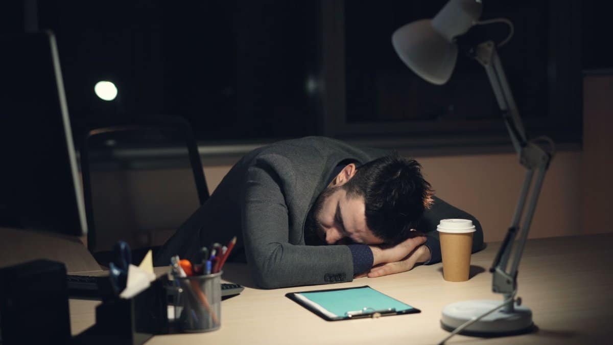 Man in office suit sleeping on desk with coffee and tablet nearby, symbolizing work fatigue.
