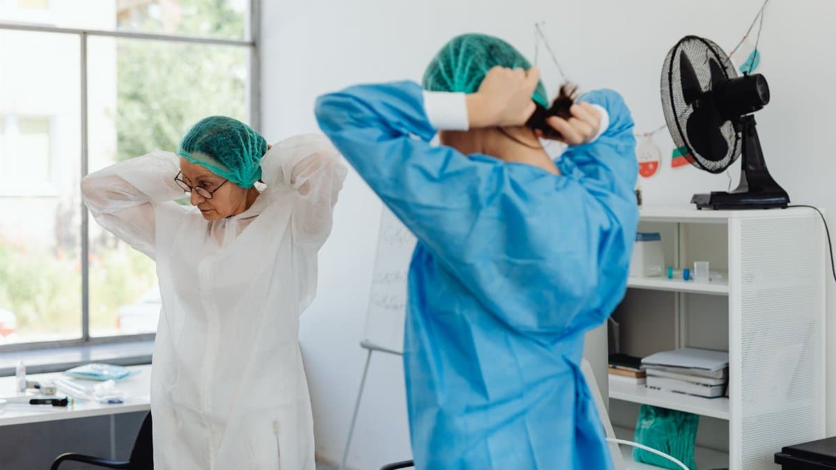 Two healthcare workers in scrubs and hair nets getting ready in a hospital room.