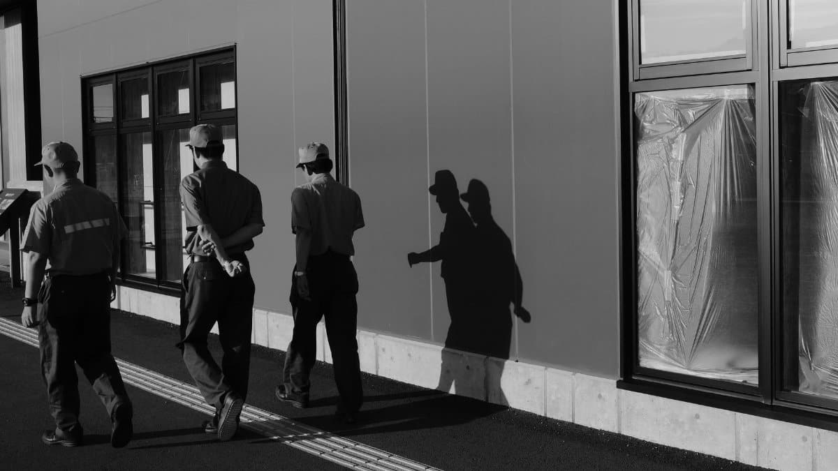 Black and white photo of workers walking along a building casting prominent shadows.