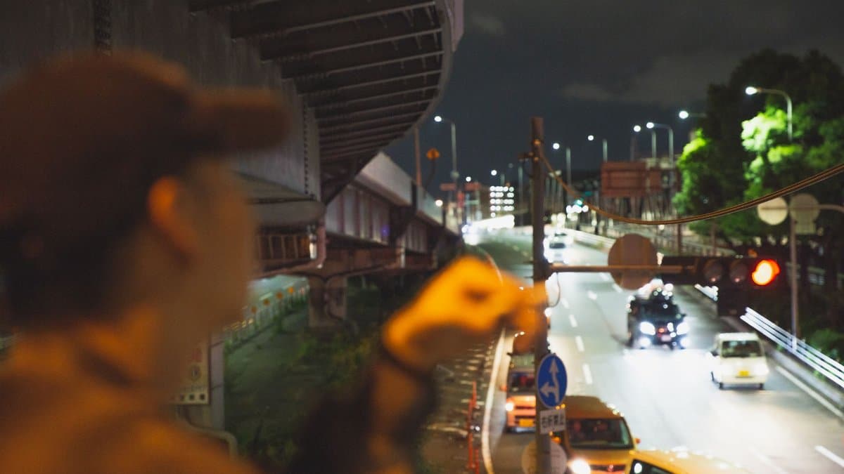Side view of male in cap with cigarette in hand standing under cars driving on road at night