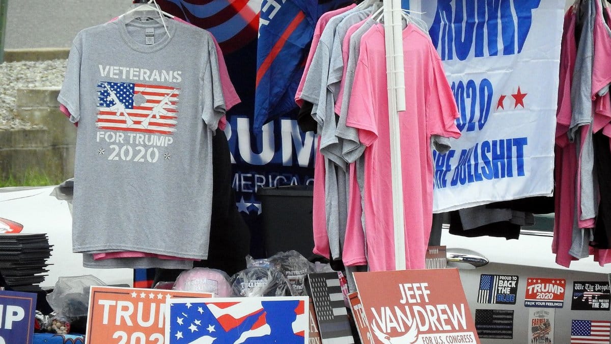 A stall displaying Trump 2020 merchandise including shirts and signs at an outdoor market.